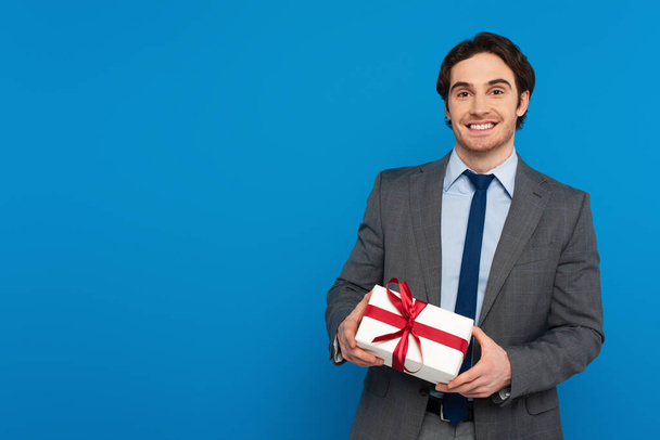 sonriente joven en blazer de moda sosteniendo caja de regalo con cinta roja aislada en azul - Foto, Imagen