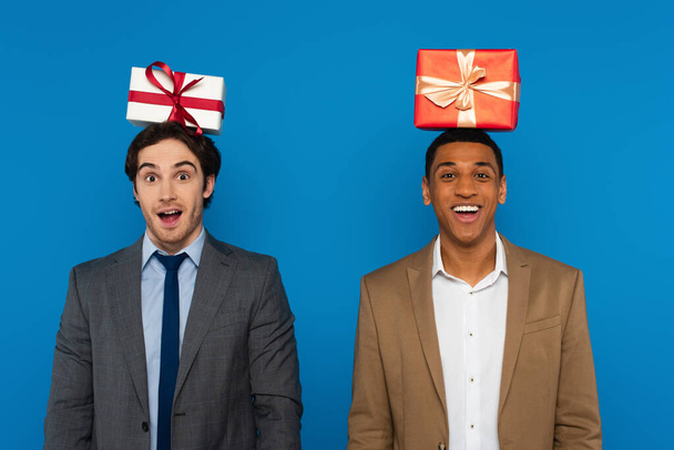 joyeux interracial amis en costumes souriant avec des boîtes-cadeaux sur les têtes isolées sur bleu - Photo, image