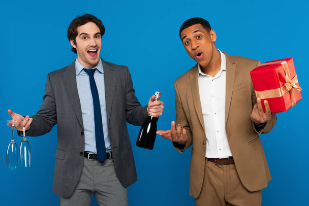 joyful interracial friends in suits holding champagne bottle, glasses and gift box isolated on blue - Foto, Imagen