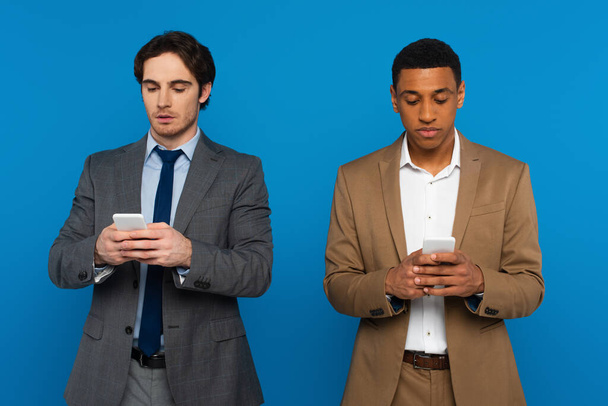 interracial men in suits watching at smartphones in hands isolated on blue - Fotó, kép