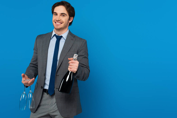 Улыбающийся молодой человек в элегантном костюме с бутылкой шампанского и стаканами, изолированными на синем баннере  - Фото, изображение