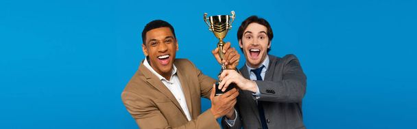 interracial přátelé v oblecích slaví úspěch s prize cup v rukou izolovaných na modré, banner  - Fotografie, Obrázek