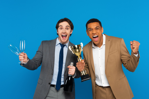 interracial friends in suits celebrating success with triumph cup, champagne and glasses in hands isolated on blue - 写真・画像
