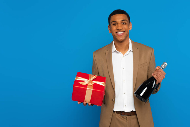 Smiling african american man in elegant suit holding champagne bottle and gift box isolated on blue - Фото, изображение