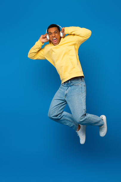 excited african american man jumping in headphones on blue background - Fotó, kép