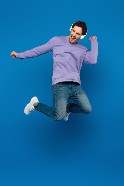 happy smiling man in violet sweatshirt enjoying music and jumping in headphones on blue background - Fotó, kép