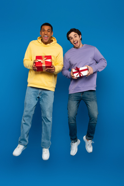 happy african american man with friend levitating with gift boxes in hands on blue background - Фото, зображення