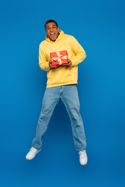 excited african american man levitating with red gift box on blue background - 写真・画像