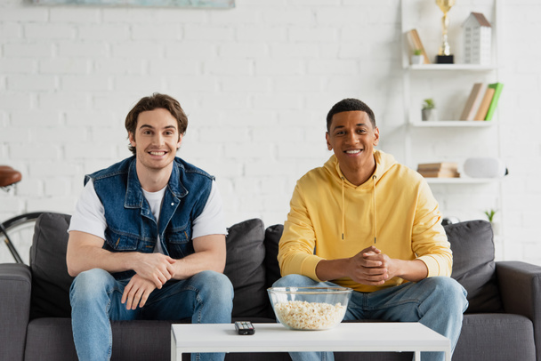smiling interracial friends sitting on couch, watching tv and eating popcorn at home - Photo, image