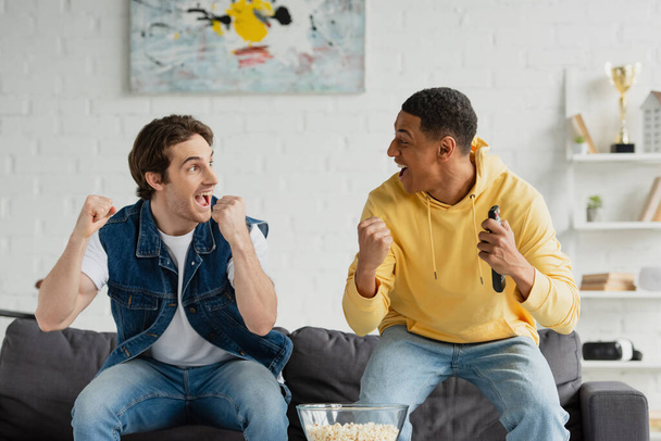 amigos interracial emocionados ver la televisión y comer palomitas de maíz en el loft moderno - Foto, imagen