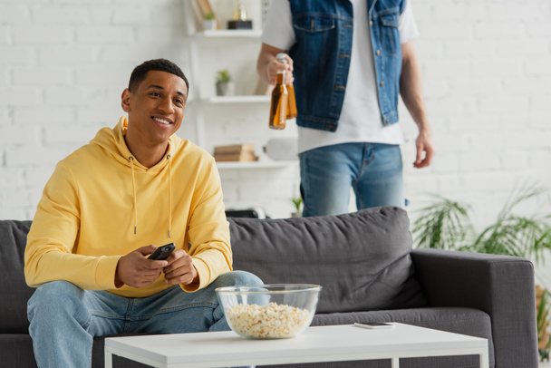 hymyilevä afrikkalainen amerikkalainen mies, jolla on tv-kaukosäädin ja jakaa olutta ystävän kanssa modernissa olohuoneessa - Valokuva, kuva