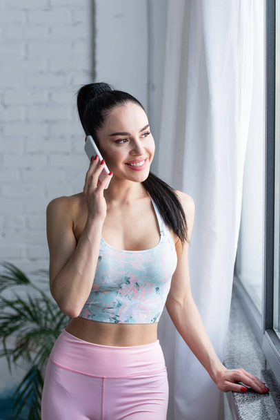 smiling woman in sports bra standing near window and talking on smartphone - 写真・画像