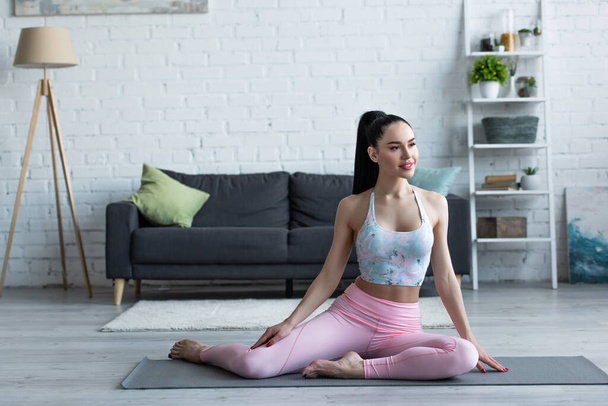 mujer sonriente mirando hacia otro lado mientras está sentada en la esterilla de yoga en casa - Foto, Imagen
