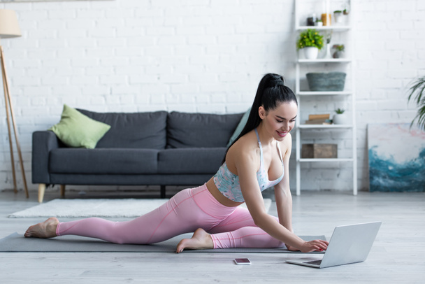 mosolygó nő nézi laptop, miközben ül jóga mat otthon - Fotó, kép