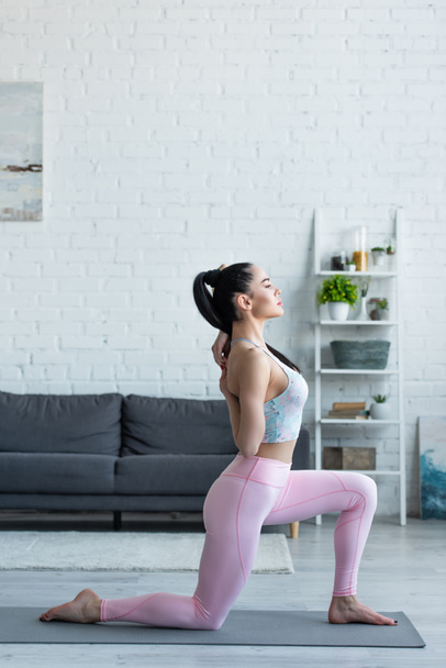 vista lateral da mulher morena praticando crescente lunge no joelho posar em casa - Foto, Imagem