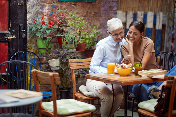 A Grandma enjoying a smartphone content with her granddaughter while they sit in a pleasant atmosphere in a bar. Leisure, bar, outdoor, friendship - Photo, image