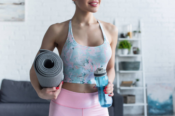 partial view of smiling woman holding fitness mat and sports bottle at home - Фото, зображення