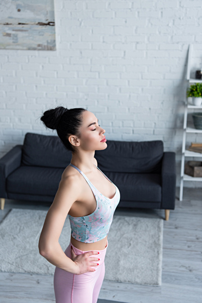 young woman in sports bra standing with hands on hips and closed eyes while training at home - Foto, afbeelding