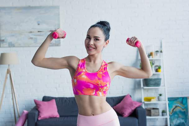 mujer deportiva sonriendo a la cámara mientras hace ejercicio con pesas en casa - Foto, Imagen