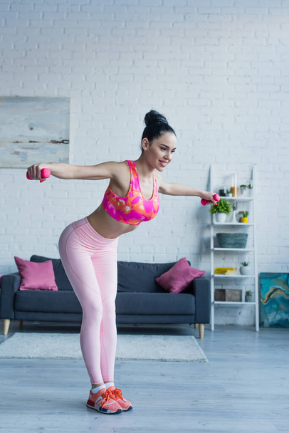 smiling woman in sports bra and leggings working out with dumbbells at home - Foto, afbeelding