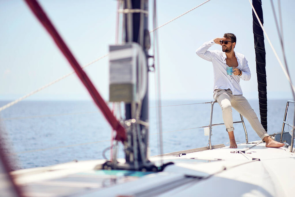 A young handsome male model is enjoying the view and coffee at a photo shooting on a yacht on a beautiful sunny day on the seaside. Summer, sea, vacation - Foto, afbeelding