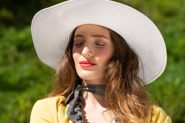 cute girl with white hat taking sun in a summer day , outside near a green grass , she has closed eyes relaxing and elegance concept - Фото, изображение