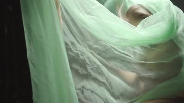 молодая женщина смотрит в камеру через зеленый шифон драпировки на сером - Кадры, видео