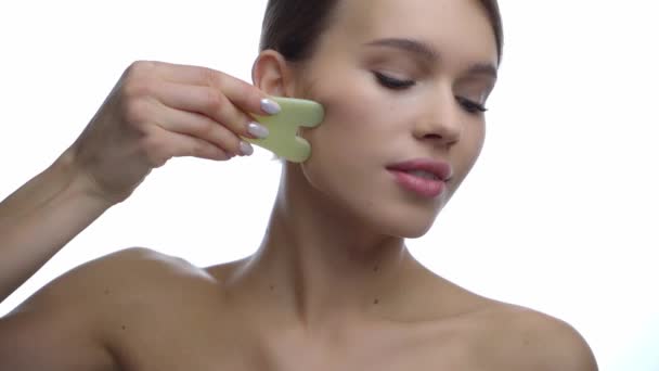 jovem mulher massageando rosto com jade raspador de pedra isolado no branco  - Filmagem, Vídeo