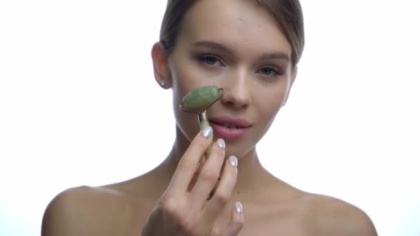 lächelnde junge Frau massiert Gesicht mit Jade-Rolle auf Weiß  - Filmmaterial, Video