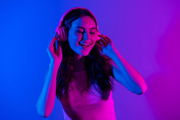Foto van jong glimlachen opgewonden mooi prachtig meisje luisteren muziek in hoofdtelefoon geïsoleerd op blauwe neon kleur achtergrond - Foto, afbeelding