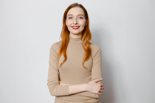 A red-haired girl on a light background crosses her arms and smiles - Foto, Bild
