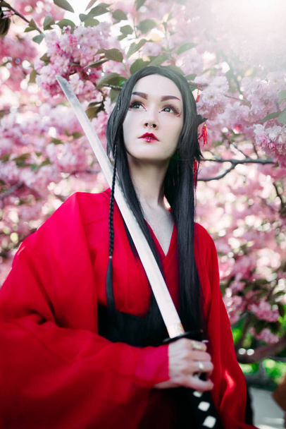 Портрет молодої жінки на зображенні гейші, одягненої в традиційне кімоно з японським самурайським мечем у руці, на тлі квітучих сакур.. - Фото, зображення