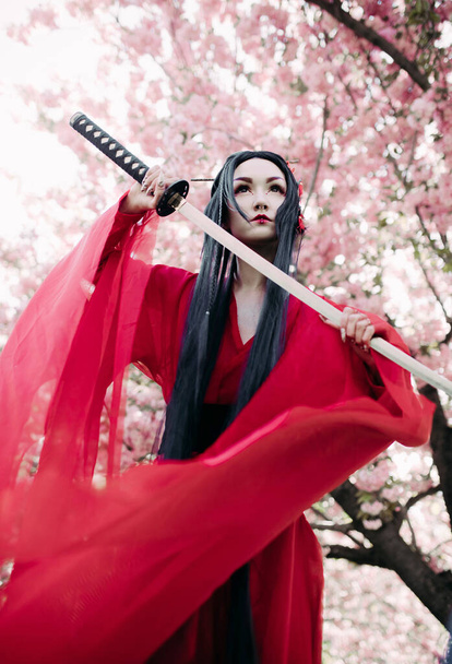 Νεαρή γυναίκα απεικονίζει γκέισα ντυμένη παραδοσιακό κιμονό με ιαπωνικό σπαθί σαμουράι στο χέρι της με φόντο ανθισμένα δέντρα σακούρα. Πίσω φανάρι. - Φωτογραφία, εικόνα