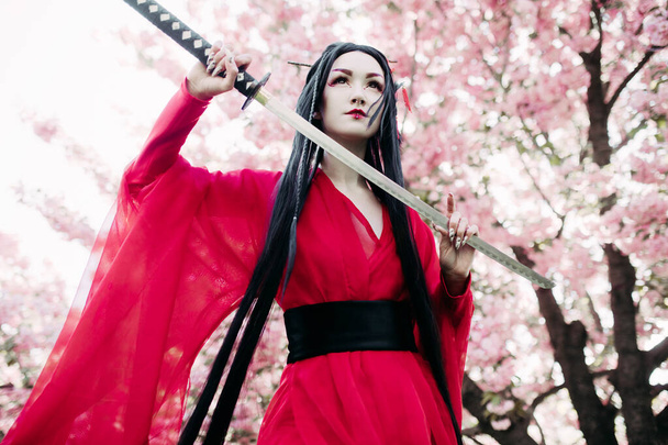 Mujer joven retrata geisha vestido kimono tradicional con espada samurai japonesa en su mano contra el fondo de los árboles de sakura florecientes. Retroiluminación. - Foto, imagen