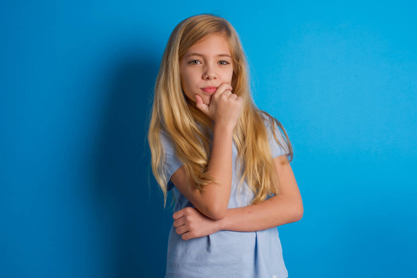 Thoughtful smiling beautiful caucasian girl wearing blue t-shirt   keeps hand under chin, looks directly at camera, listens something with interest. Youth concept. - Foto, Bild