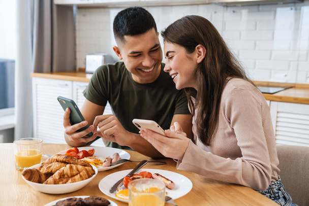 Portrait of smiling couple using smartphones and talking while having breakfast in cozy kitchen at home - Foto, Imagem