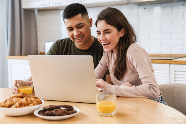 Smiling happy couple enjoying breakfast in the kitchen looking at laptop screen making video call - Φωτογραφία, εικόνα
