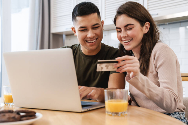 Portrait of a cheerful couple shopping online with laptop computer while sitting at the table in a kitchen at home, woman holding credit card - 写真・画像