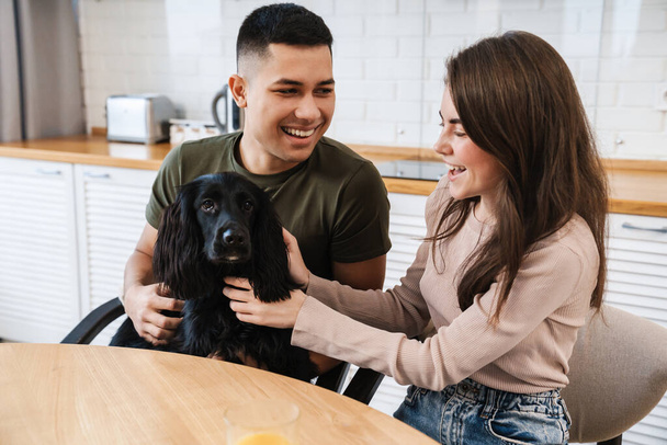 Cheerful lovely couple with their prt dog sitting in a kitchen, playing - Photo, Image