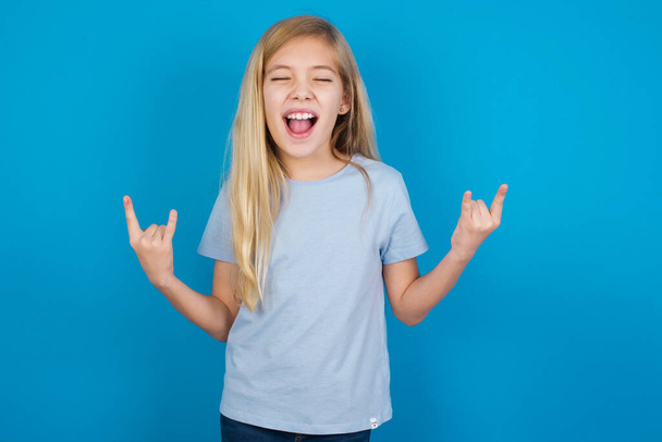 Рожден, чтобы потрясти этот мир. Радостная красивая кавказская девушка в голубой футболке, громко кричащая и показывающая с поднятыми руками рога или жест камня. - Фото, изображение