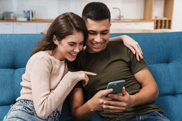 Smiling happy couple using mobile phone while sitting on a couch at home - Foto, Imagem