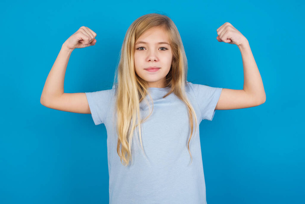Waist up shot of beautiful caucasian girl wearing t-shirt   raises arms to show muscles feels confident in victory, looks strong and independent, smiles positively at camera. Sport concept. - Photo, Image
