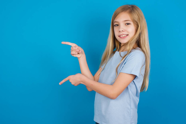 bella caucasica bambina con lunghi capelli biondi indossando t-shirt che punta con le dita su sfondo blu - Foto, immagini