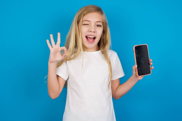Excited  beautiful caucasian girl wearing t-shirt    showing smartphone blank screen, blinking eye and doing ok sign with hand.  Advertisement concept. - Foto, immagini