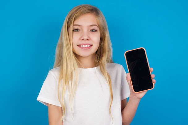 Lächelnde schöne kaukasische Mädchen im T-Shirt, das den leeren Telefonbildschirm zeigt. Werbe- und Kommunikationskonzept. - Foto, Bild