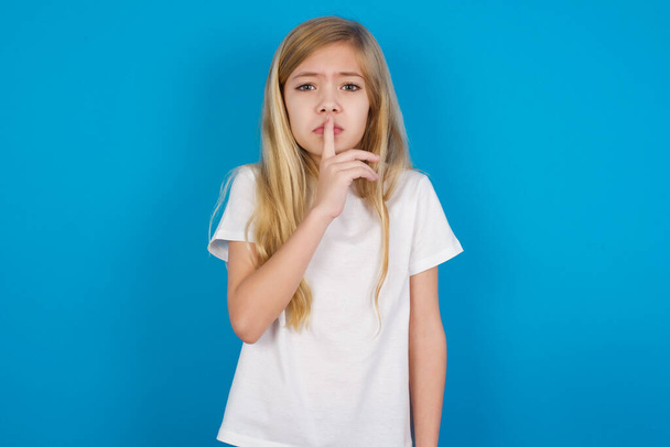 beautiful caucasian girl wearing t-shirt   makes silence gesture, keeps index finger to lips makes hush sign. Asks not to share secret. - Foto, Bild