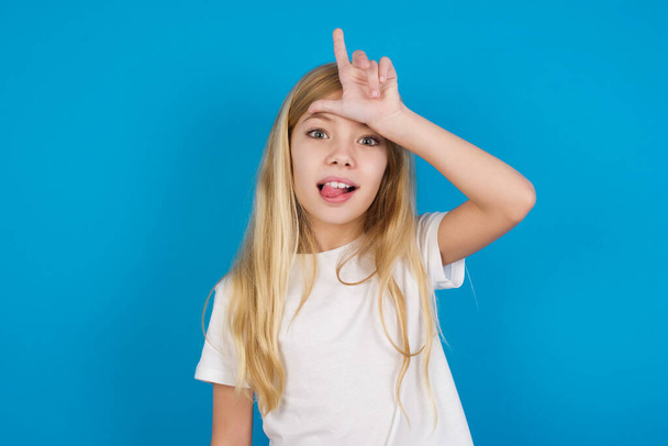  gyönyörű kaukázusi lány visel póló gesztusok ujj a homlokán teszi vesztes gesztus viccet emberek mutatja nyelv - Fotó, kép