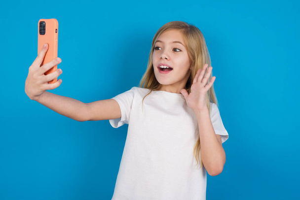  όμορφη καυκάσια κοπέλα φορώντας t-shirt κατέχει σύγχρονο κινητό τηλέφωνο και κάνει video call παλάμη κύματα σε χειρονομία γεια. Η έννοια της σύγχρονης τεχνολογίας - Φωτογραφία, εικόνα