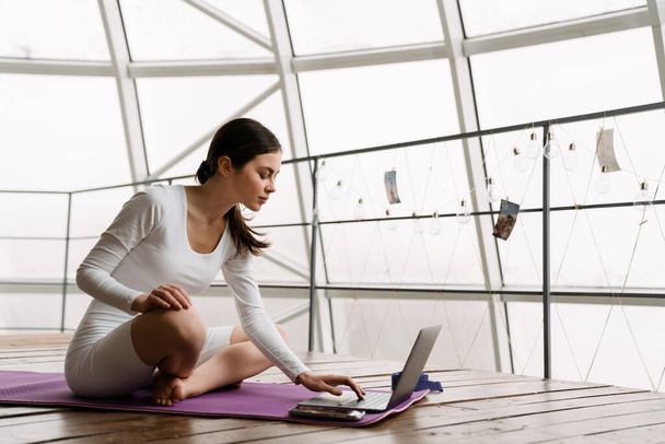 Jeune femme blanche utilisant un ordinateur portable pendant la pratique du yoga à l'intérieur - Photo, image