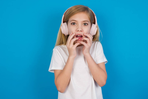 Wstrząśnięty piękny kaukaski dziewczyna noszenie t-shirt gapi się strach na aparat utrzymuje usta szeroko otwarte nosi bezprzewodowe słuchawki stereo na uszach - Zdjęcie, obraz